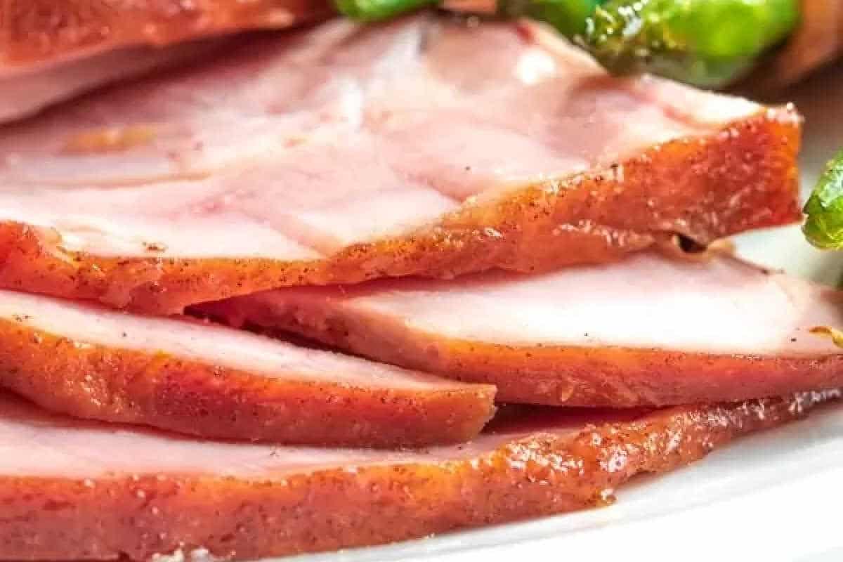 Close-up of several slices of cooked ham with a brown outer edge, arranged on a plate with green vegetables in the background.