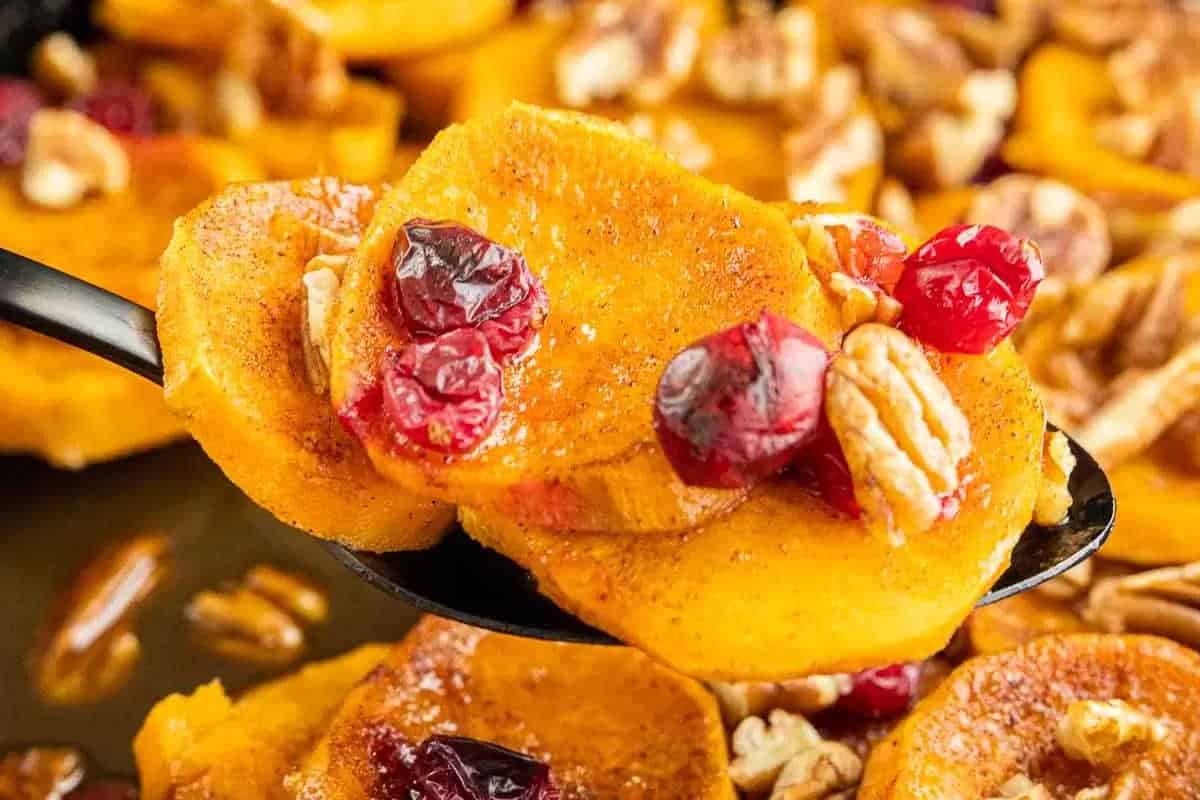 Close-up of roasted sweet potato slices topped with dried cranberries and pecans on a spoon, with more on a tray in the background—the perfect plate for your Christmas sides lineup.