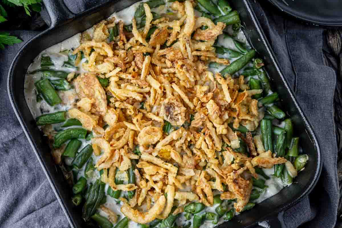 A casserole dish filled with green bean casserole topped with crispy fried onions, set on a dark cloth—an essential addition to the ultimate lineup of Christmas sides for the perfect plate.