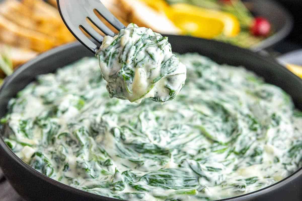 A fork lifts a serving of creamy spinach dip from a black bowl, creating the perfect plate for your Christmas sides and the ultimate lineup of holiday appetizers.