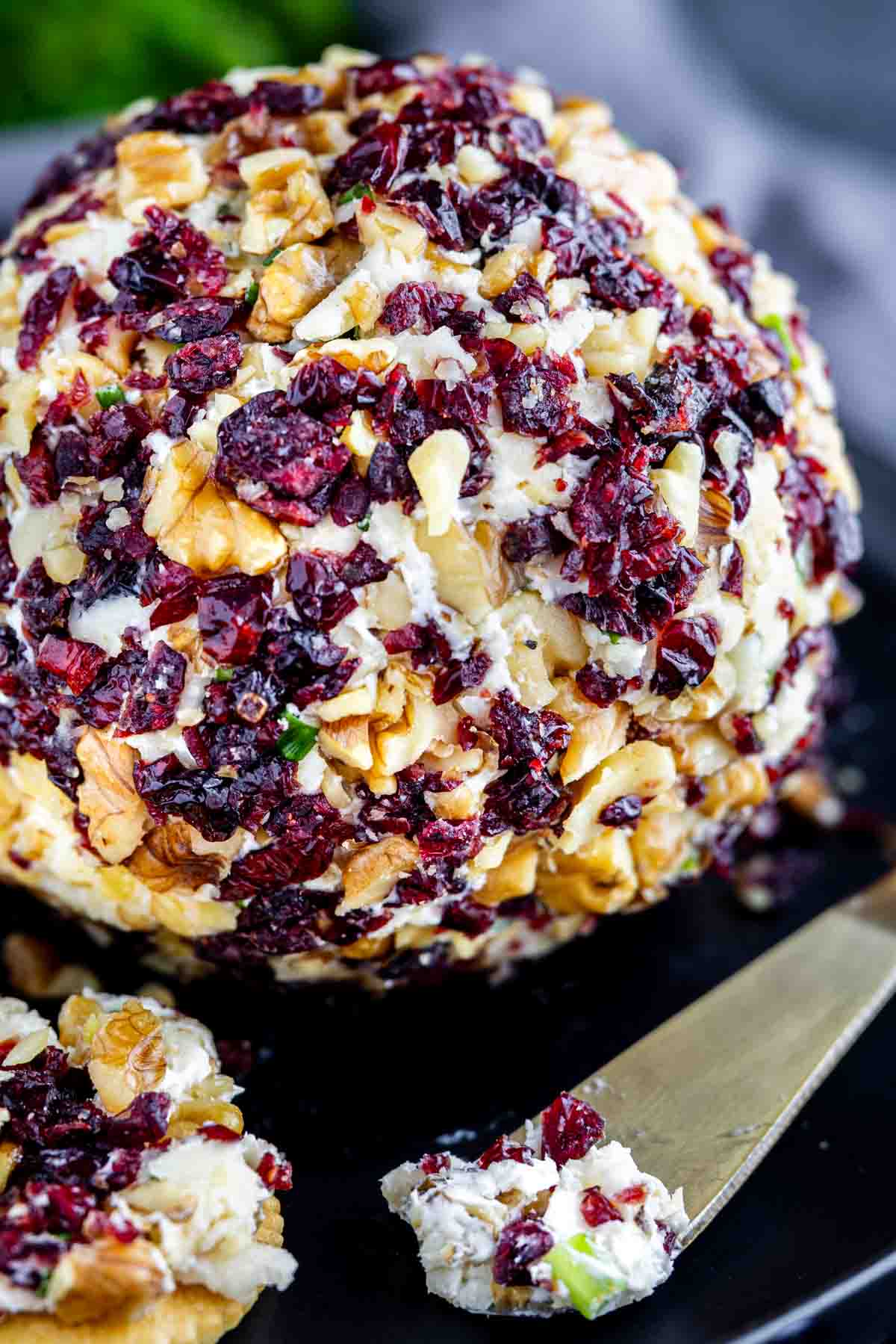 A close-up of a cheese ball with cranberries and chopped nuts, with a knife spreading the festive cheeseball recipe onto a cracker.
