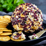 A cheese ball coated with chopped nuts and dried cranberries is served on a black plate with round crackers and a spreading knife.