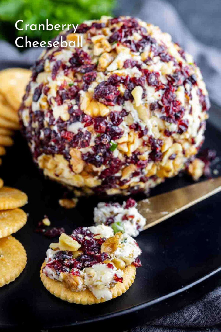 A cranberry cheeseball coated with chopped walnuts and cranberries, served with round crackers on a black plate.