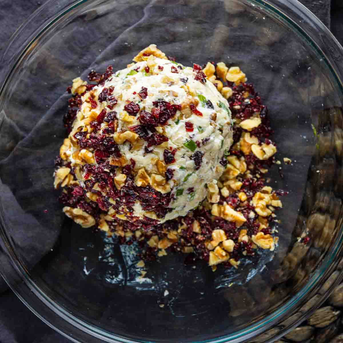 A cranberry cheeseball coated with chopped dried cranberries and walnuts sits in a clear glass bowl on a dark cloth.