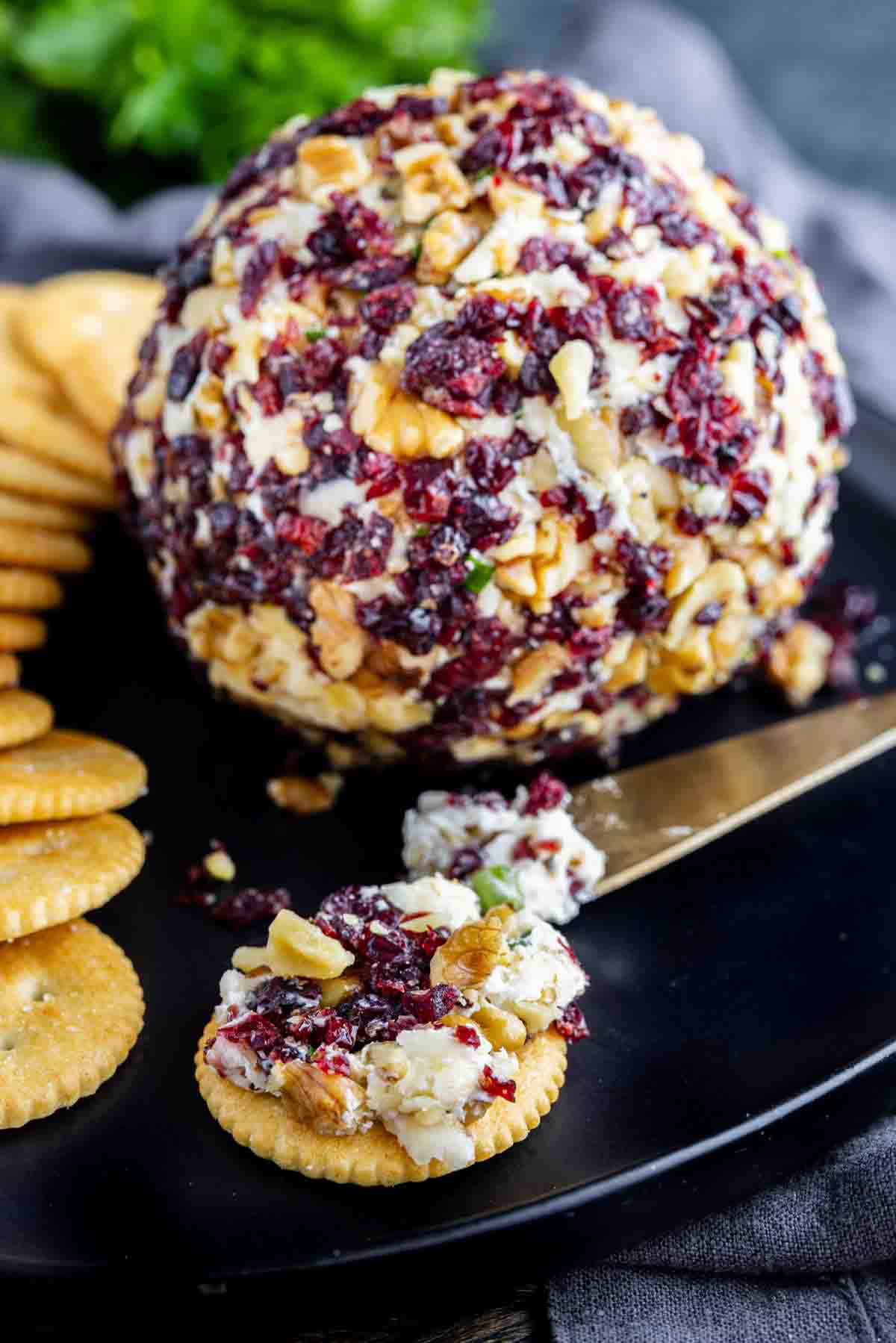 A cheese ball with cranberries and chopped nuts is served on a black plate with crackers and a cheese knife—a festive cheeseball recipe for any occasion.
