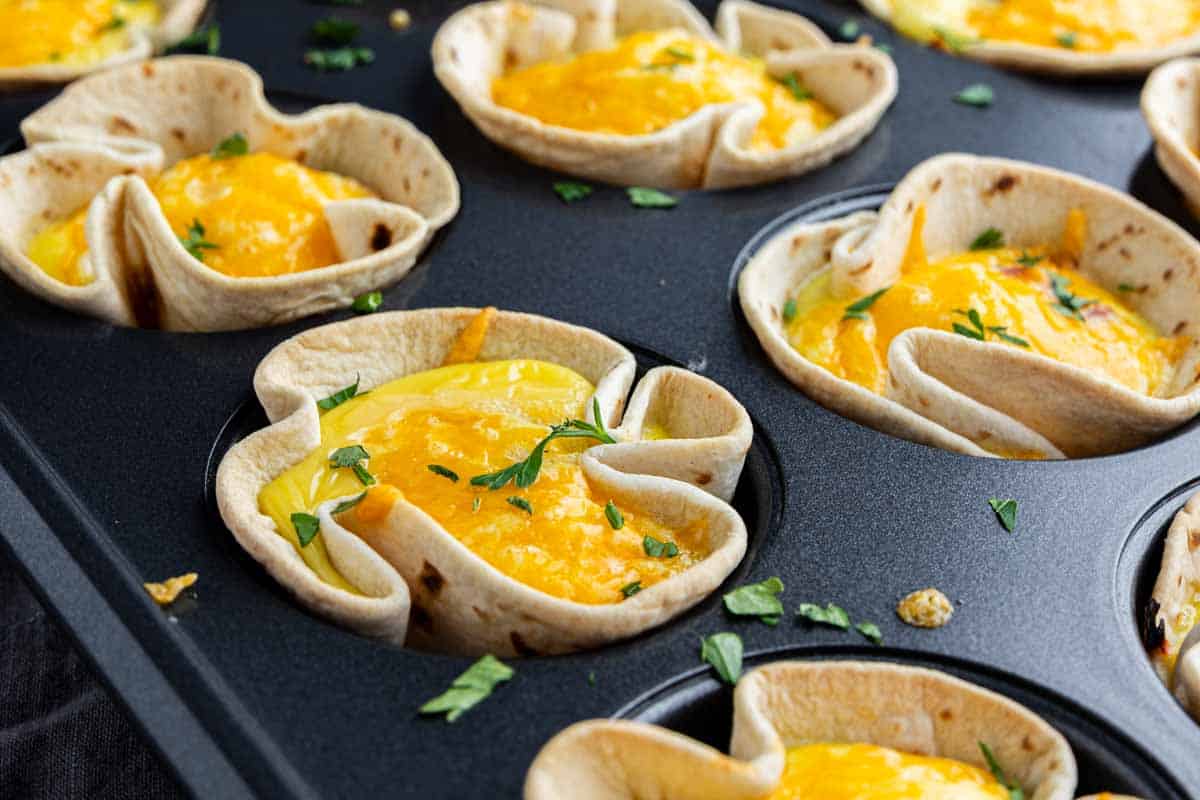 Muffin tin filled with tortilla cups containing baked eggs, melted cheese, and chopped herbs.