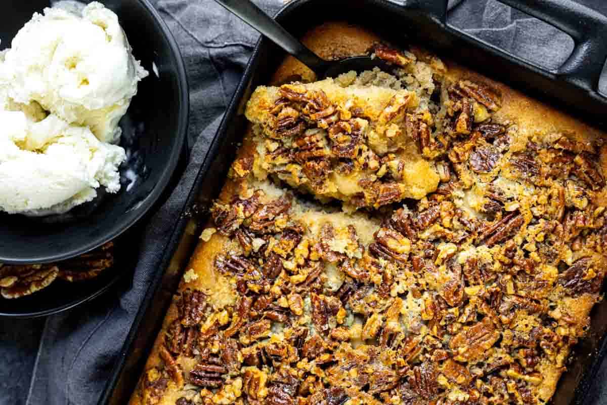 A baking dish filled with pecan cobbler sits next to a bowl of vanilla ice cream. A serving has been scooped from the cobbler.