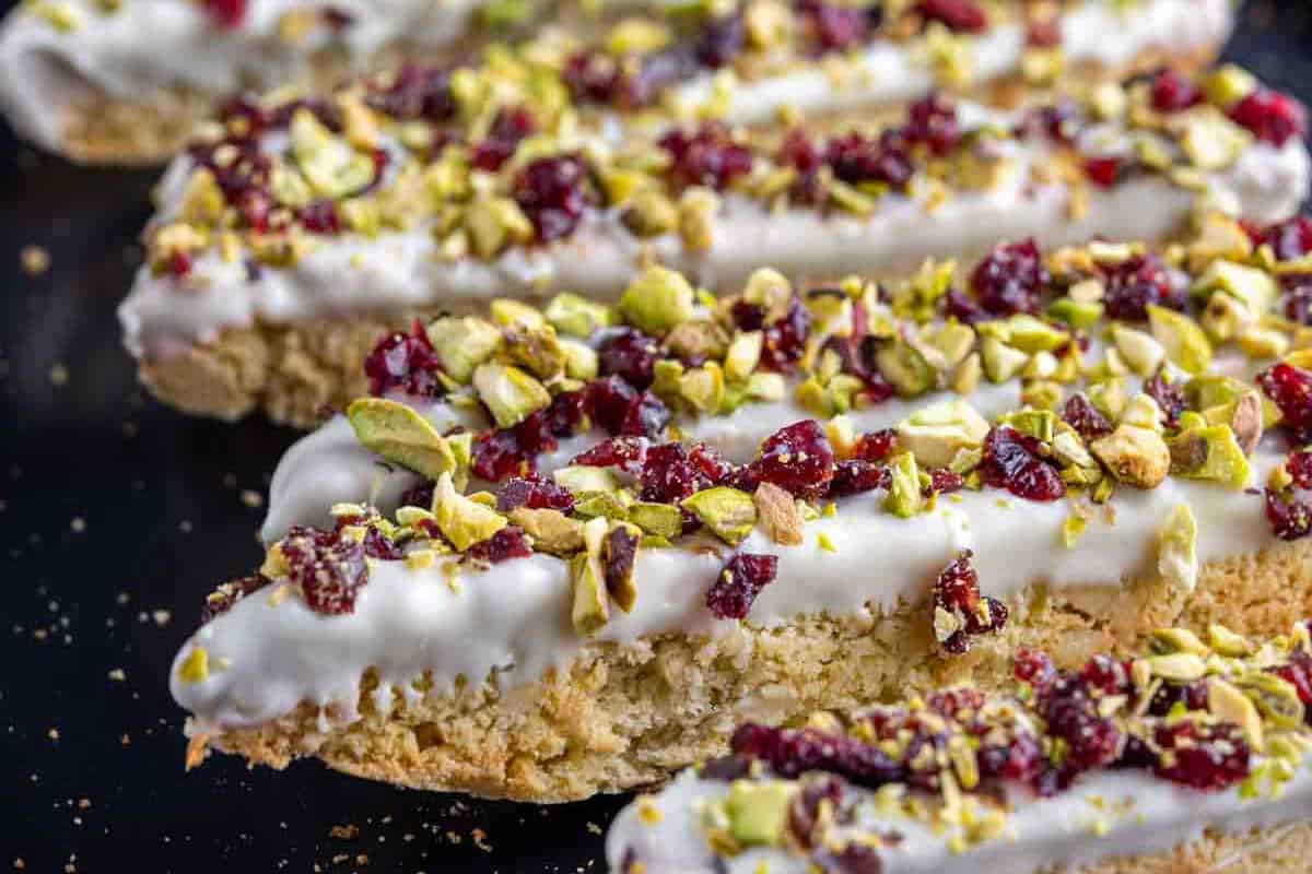 Close-up of biscotti cookies dipped in white chocolate and topped with chopped pistachios and dried cranberries on a dark surface.