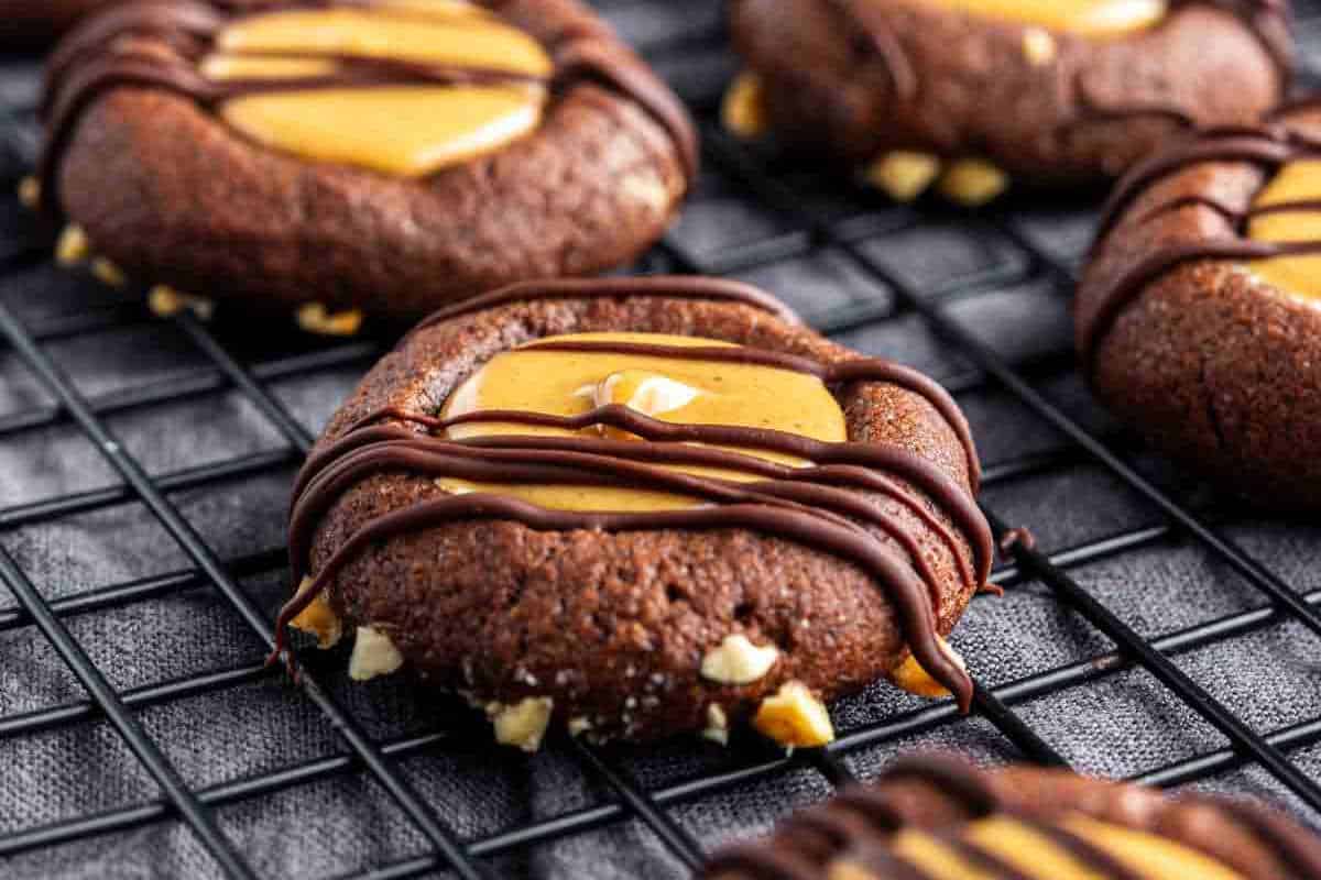 Chocolate cookies with a peanut butter center and chocolate drizzle, cooling on a wire rack. Pieces of chopped nuts are visible around the cookie edges.