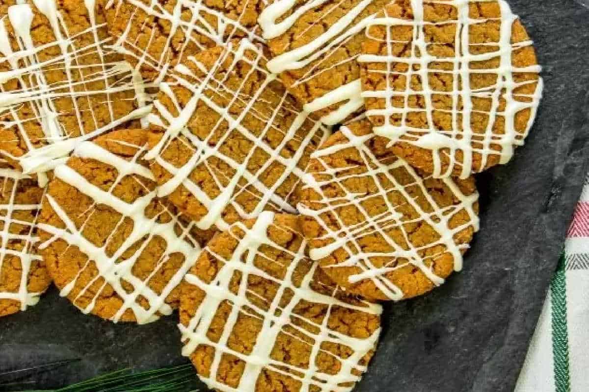 A group of round, brown cookies with a white icing drizzle arranged on a dark slate surface.