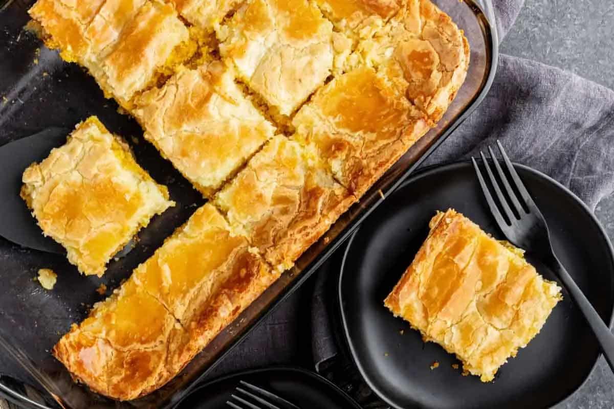 A glass baking dish with sliced gooey butter cake, one piece served on a black plate with a fork on the side.