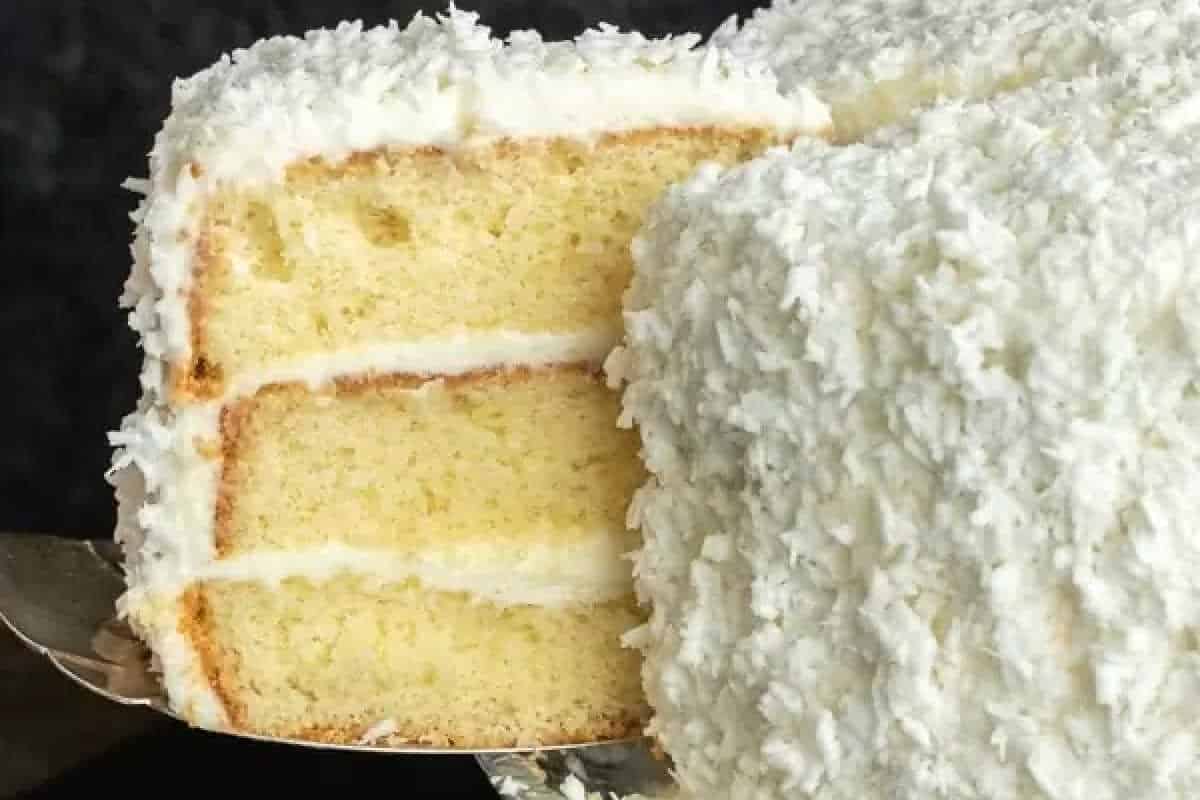 A three-layer vanilla cake with white frosting and shredded coconut on the outside, with one slice being served.