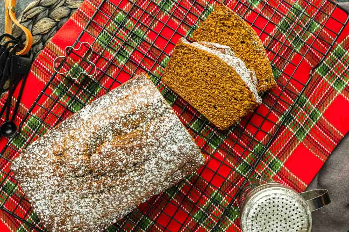 A loaf of bread dusted with powdered sugar on a cooling rack, with two slices cut, placed on a red plaid cloth. A sifter, cookie cutter, and measuring spoons are nearby.