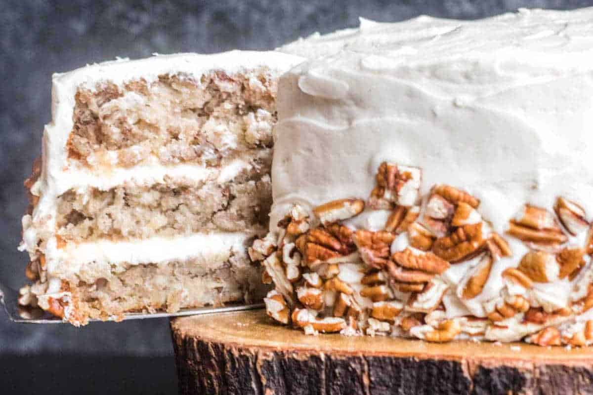 A three-layer cake with white frosting and chopped pecans on the side, displayed on a wooden stand with one slice being removed.