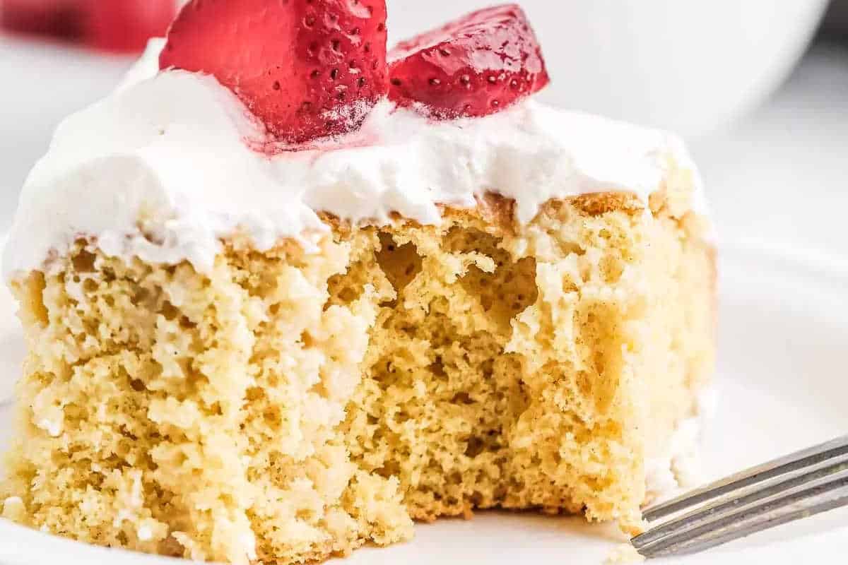 A slice of yellow sponge cake with white frosting and sliced strawberries on top, with a bite taken out and a fork beside it.