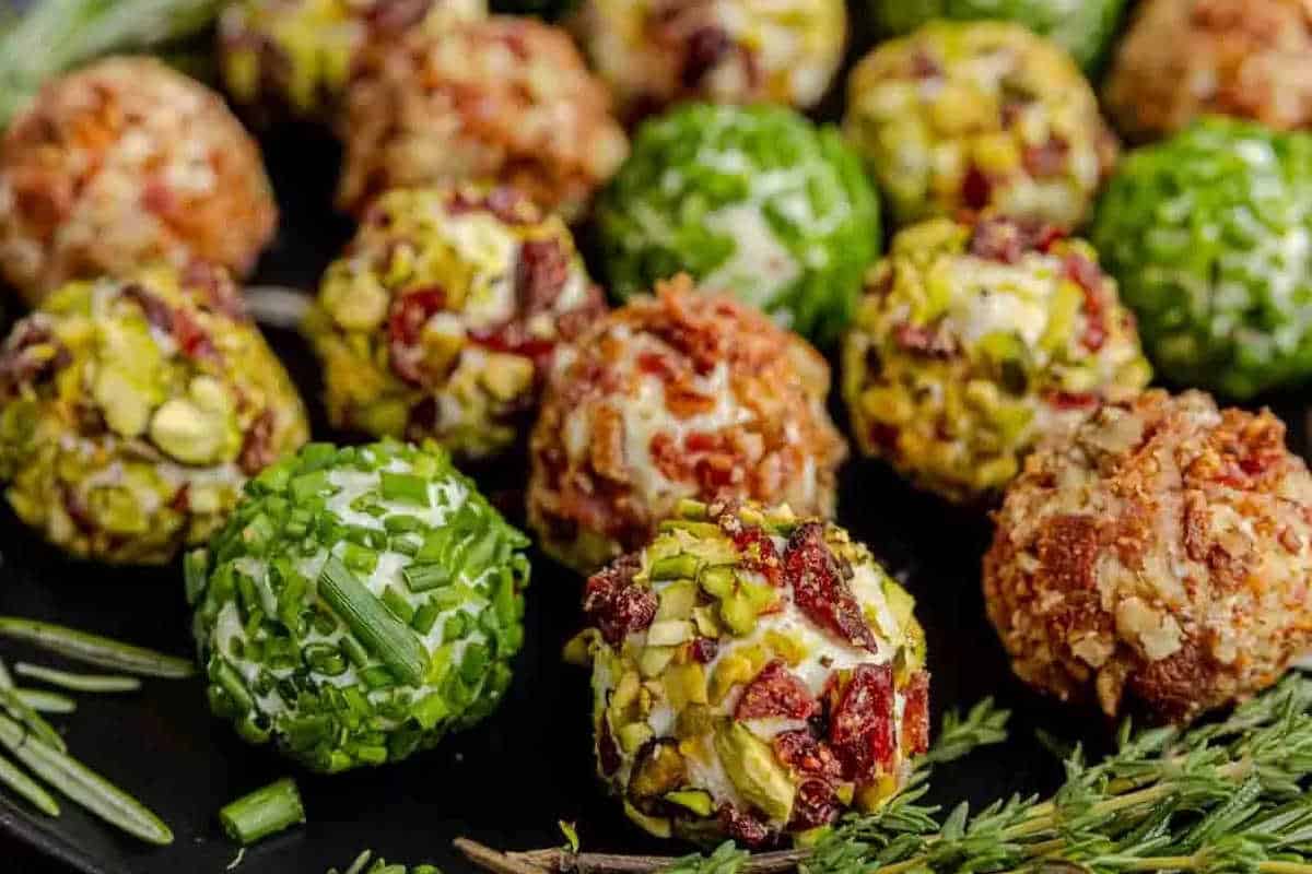 Assorted cheese balls coated in chopped herbs, nuts, and bacon bits are arranged on a dark platter with sprigs of fresh rosemary and thyme.