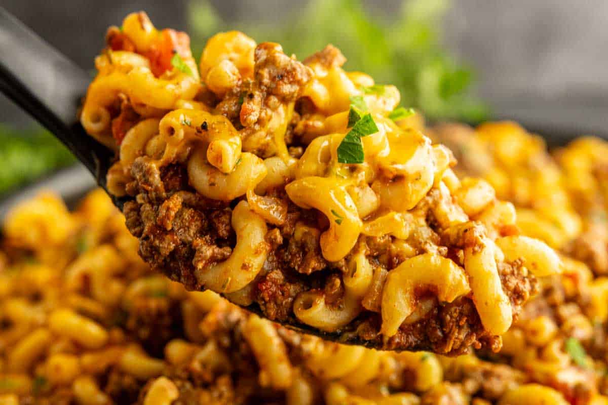 A close-up of a spoonful of macaroni pasta mixed with ground beef, tomato sauce, and melted cheese, garnished with chopped herbs.