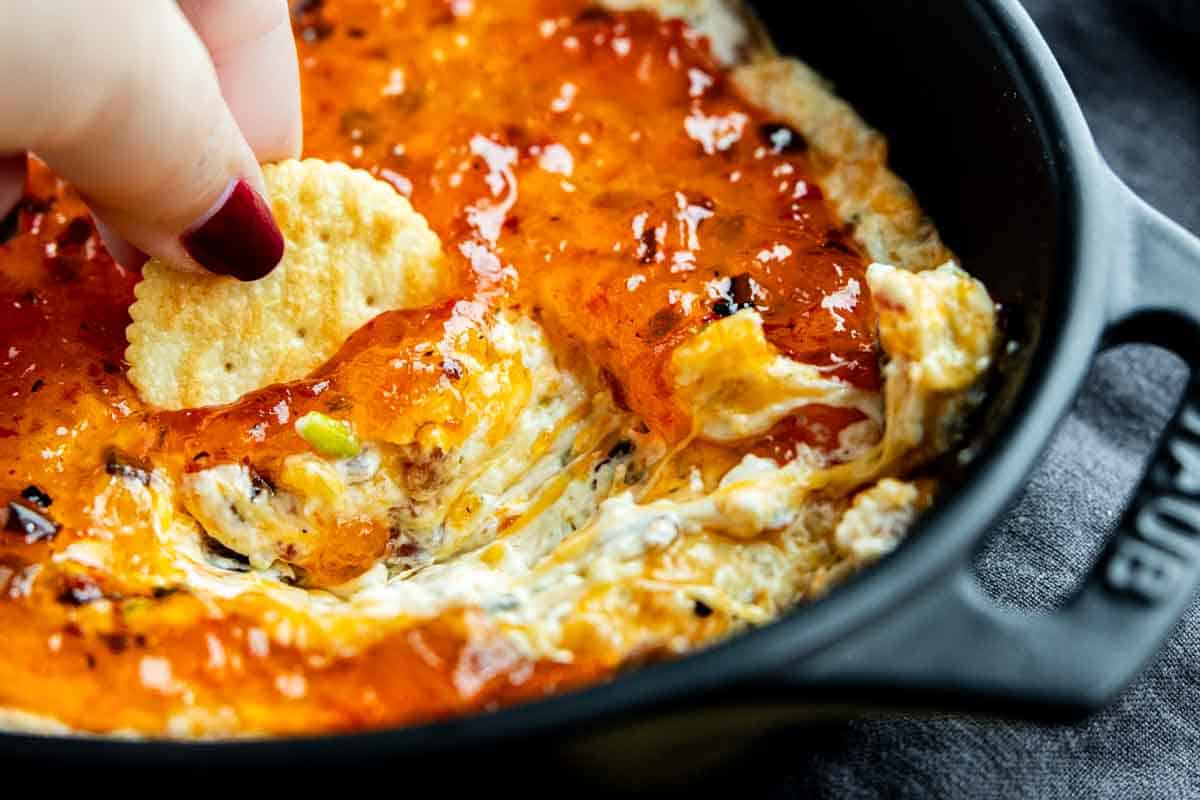 A hand dipping a round cracker into a creamy cheese dip topped with a layer of orange-red jelly in a black dish.
