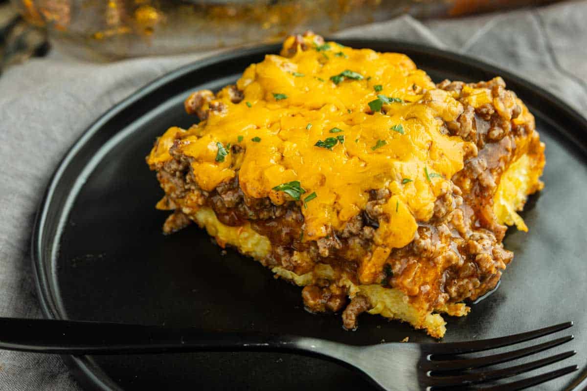 A slice of casserole topped with melted cheddar cheese and ground beef sits on a black plate with a black fork.
