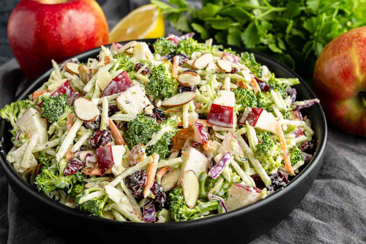 A bowl of salad with broccoli, apple chunks, shredded carrots, sliced almonds, and cranberries, with apples and herbs in the background.