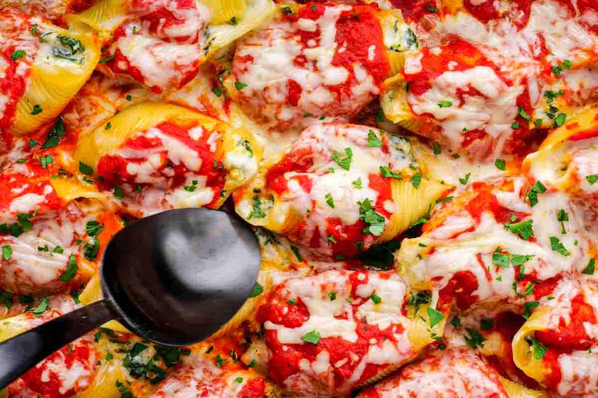 Stuffed pasta shells topped with tomato sauce, melted cheese, and chopped herbs, with a black serving spoon resting on the dish.