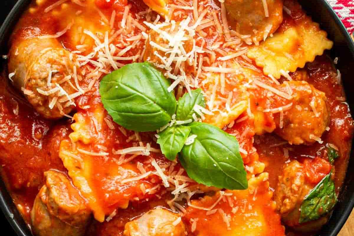 A bowl of ravioli in tomato sauce with meatballs, topped with grated cheese and fresh basil leaves.