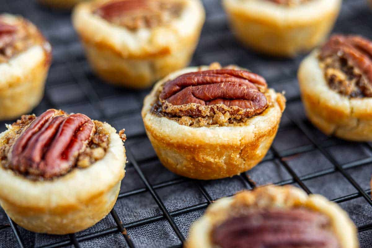Mini pecan tarts with golden crusts and whole pecans on top are arranged on a cooling rack.