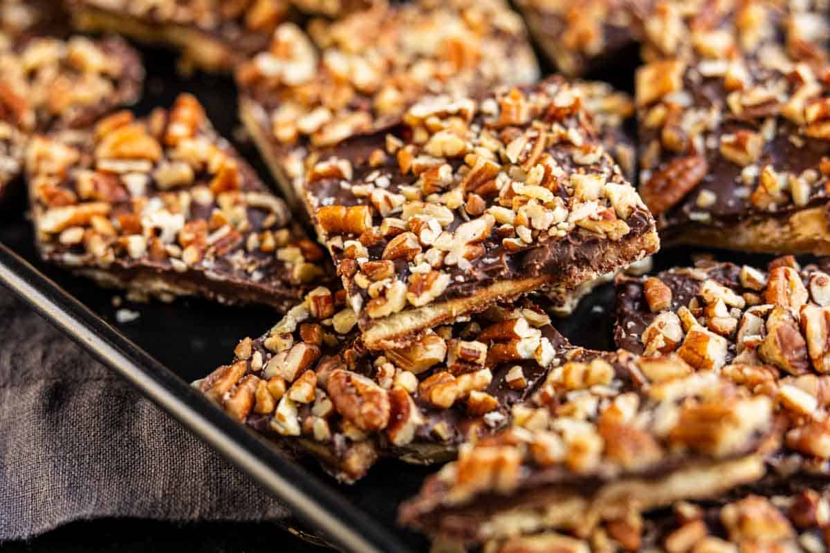 A close-up of toffee squares topped with chopped pecans and a layer of chocolate, arranged in a slightly overlapping pattern on a dark tray.