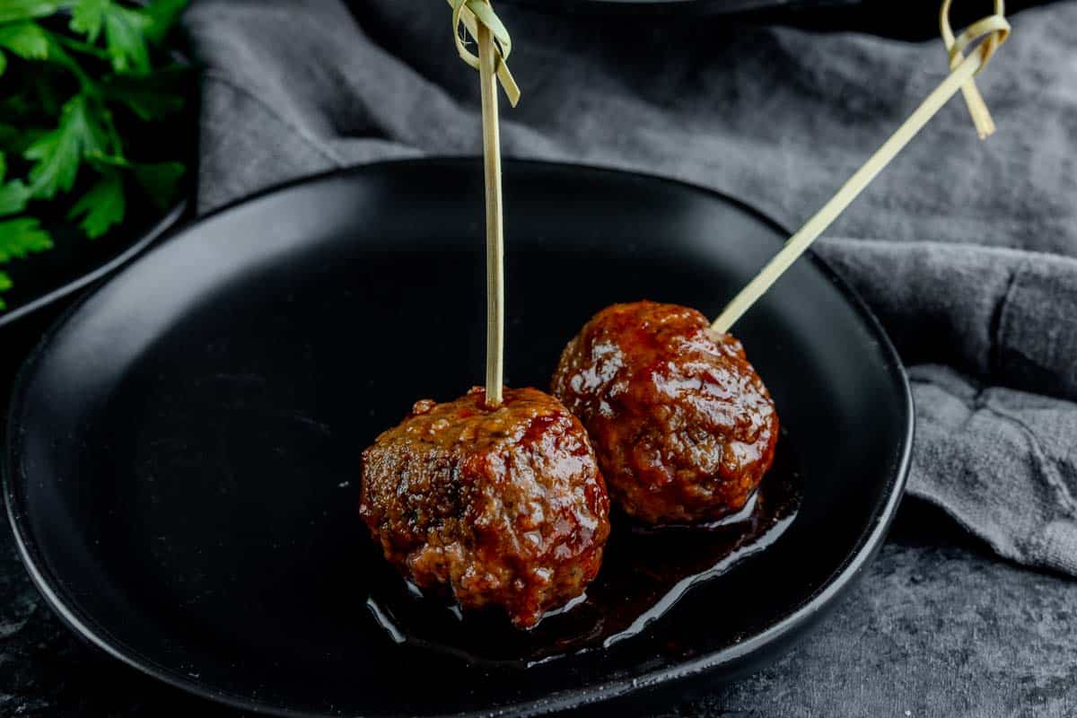 Two glazed meatballs with toothpicks are served on a black plate, with a dark cloth and parsley in the background.