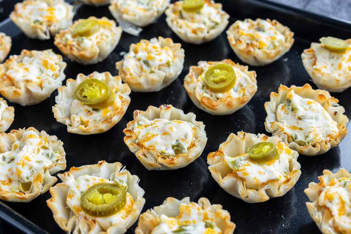 Mini phyllo cups filled with creamy cheese mixture, some topped with sliced jalapeños, make perfect last-minute appetizers for Christmas parties, arranged beautifully on a black baking sheet.