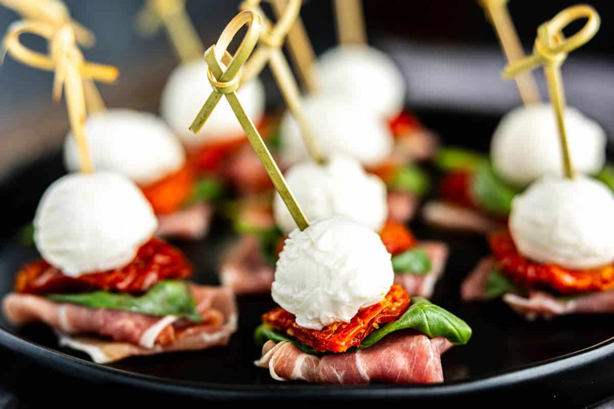 These no stress skewers with mozzarella balls, sun-dried tomatoes, basil leaves, and prosciutto are perfect as last-minute appetizers for Christmas parties, beautifully arranged on a black plate.