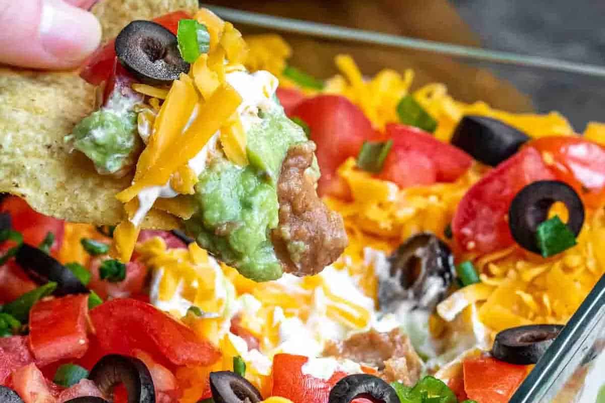 A close-up of a tortilla chip scooping seven-layer dip—perfect for Christmas parties or last-minute appetizers—with refried beans, guacamole, sour cream, shredded cheese, tomatoes, olives, and green onions.