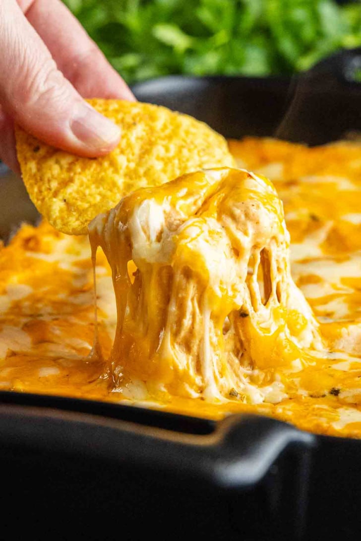 A hand dips a tortilla chip into a cheesy, melted chicken enchilada dip in a black dish, with gooey cheese stretching between the chip and the savory game day dip.