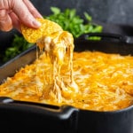 A hand dips a tortilla chip into a cheesy baked chicken dip, pulling up melted cheese from a black baking dish with green herbs in the background—perfect as a game day dip.