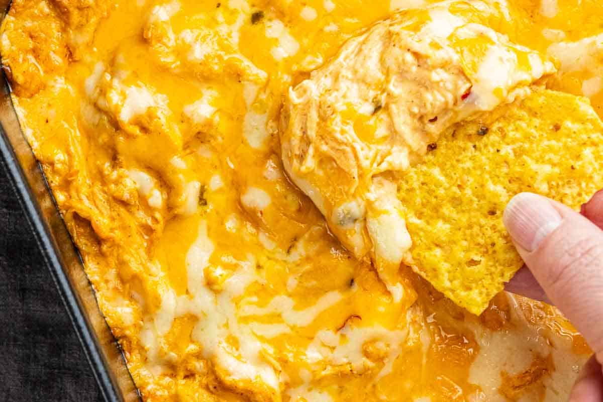 A hand dips a tortilla chip into a cheesy baked chicken enchilada dip in a rectangular glass dish.