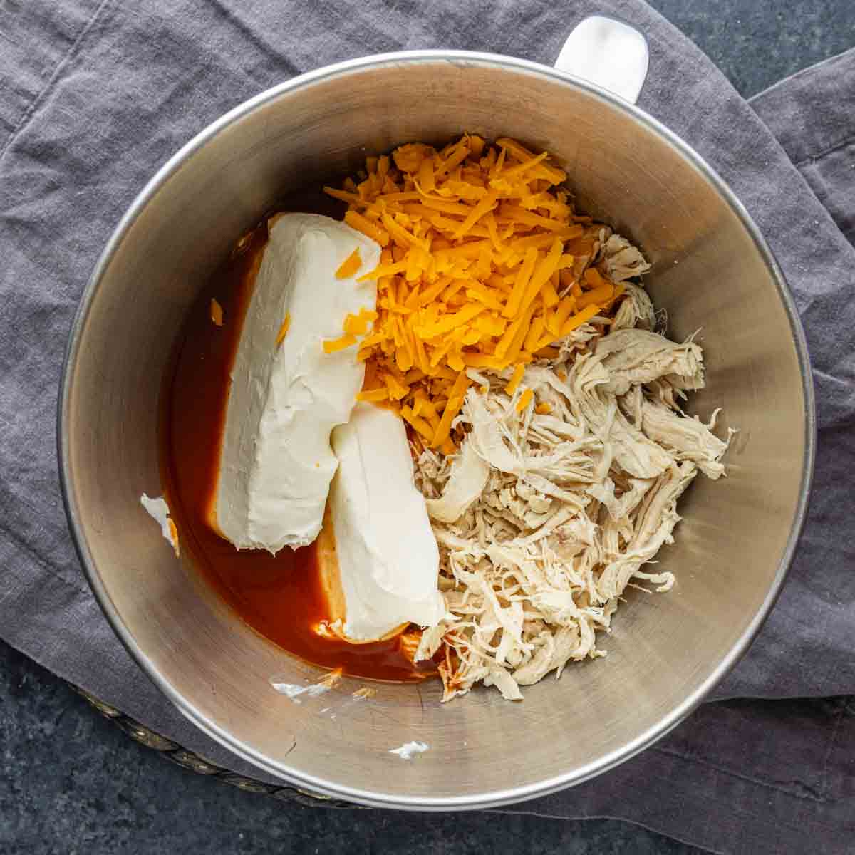 A metal mixing bowl containing shredded chicken, cheddar cheese, cream cheese blocks, and buffalo sauce sits on a gray cloth—perfect for a zesty baked chicken dip or as a crowd-pleasing game day dip.