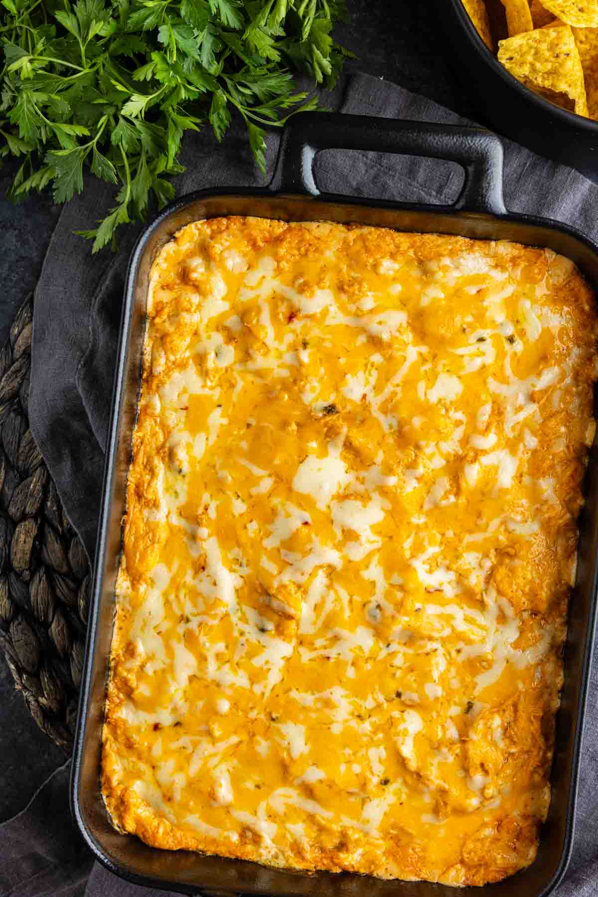 A baking dish filled with golden, melted cheese-topped chicken enchilada dip sits on a dark surface next to fresh parsley and a bowl of tortilla chips.