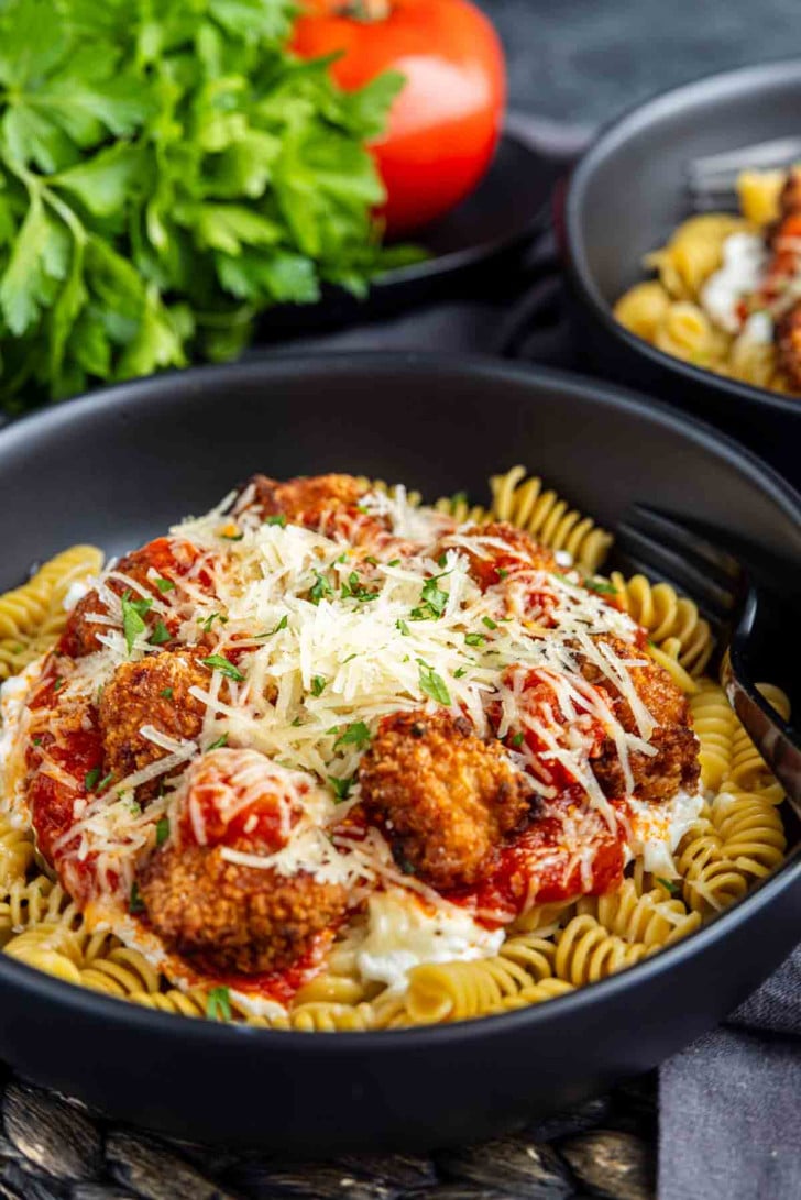 A black bowl filled with rotini pasta, topped with breaded meatballs, tomato sauce, melted cheese, and shredded parmesan, with fresh parsley and a tomato in the background.