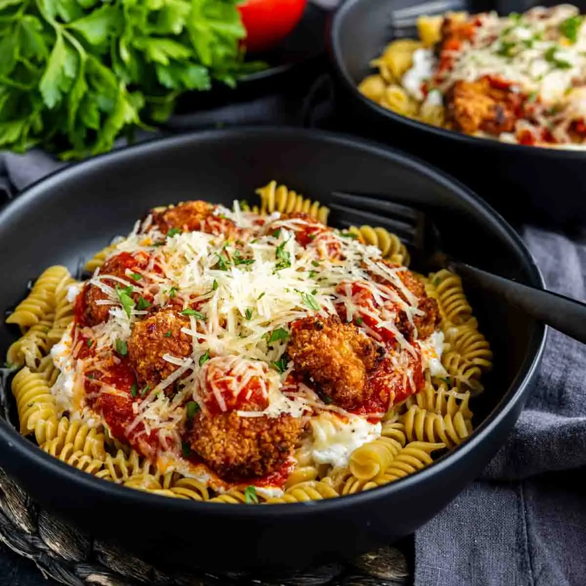 A black bowl filled with rotini pasta, topped with high protein chicken parmesan meatballs, marinara sauce, grated cheese, and herbs. A fork rests in the bowl and fresh parsley is nearby—an easy high protein lunch option.