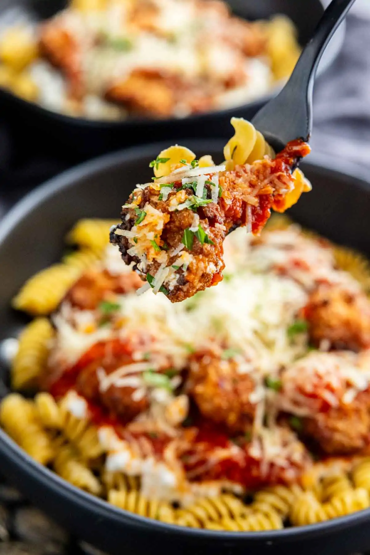 A close-up of a fork holding a bite of pasta with tomato sauce, grated cheese, and meatball over a bowl of rotini pasta topped with sauce and cheese—an easy high protein dinner perfect for busy nights.