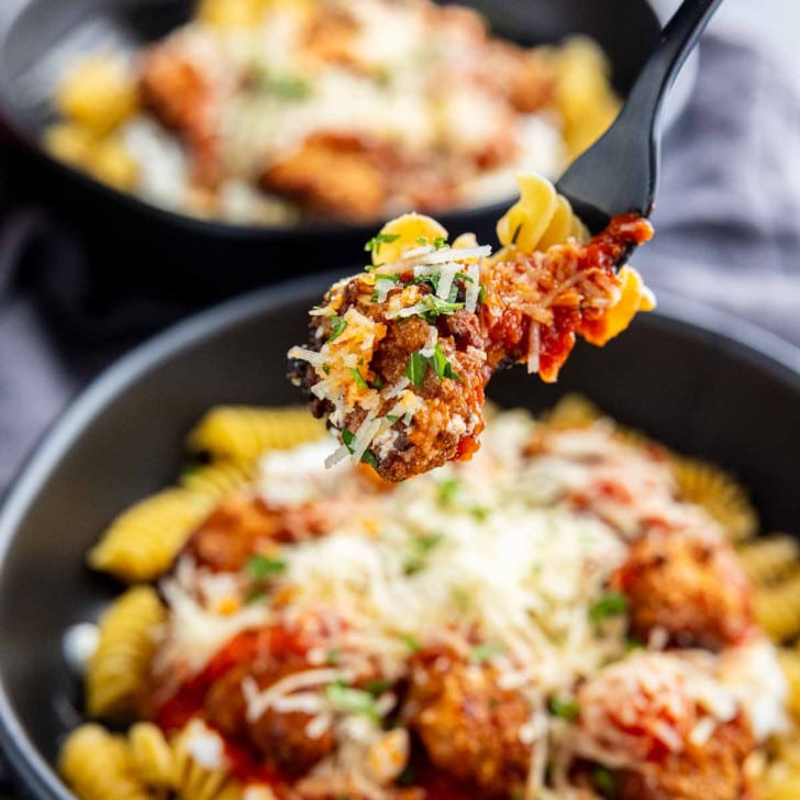 A close-up of a fork holding pasta, meatball, sauce, and cheese above a bowl filled with rotini pasta, meatballs, tomato sauce, and grated cheese.