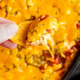 A hand holds a corn chip topped with melted cheese and dip, with more cheesy dip visible in the background.