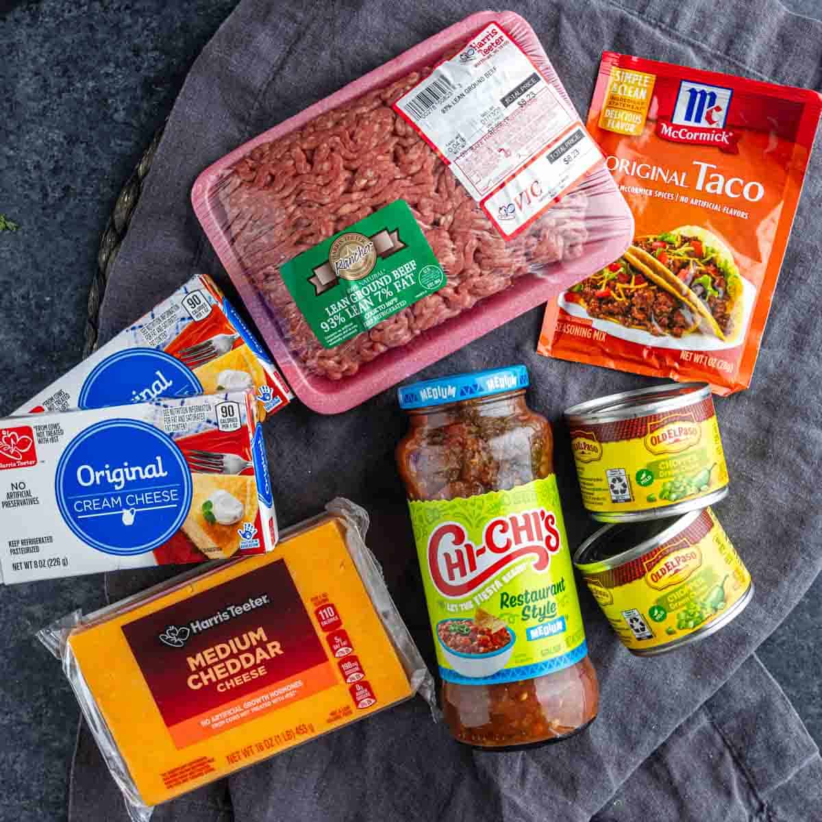 Top-down view of easy taco dip ingredients: ground beef, taco seasoning, two cream cheese packs, cheddar cheese, salsa, and two cans of diced green chiles on a dark surface.