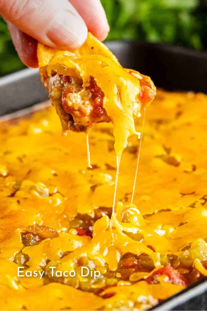 A hand dips a tortilla chip into a cheesy taco dip, lifting a portion with melted cheese stretching from the dish.