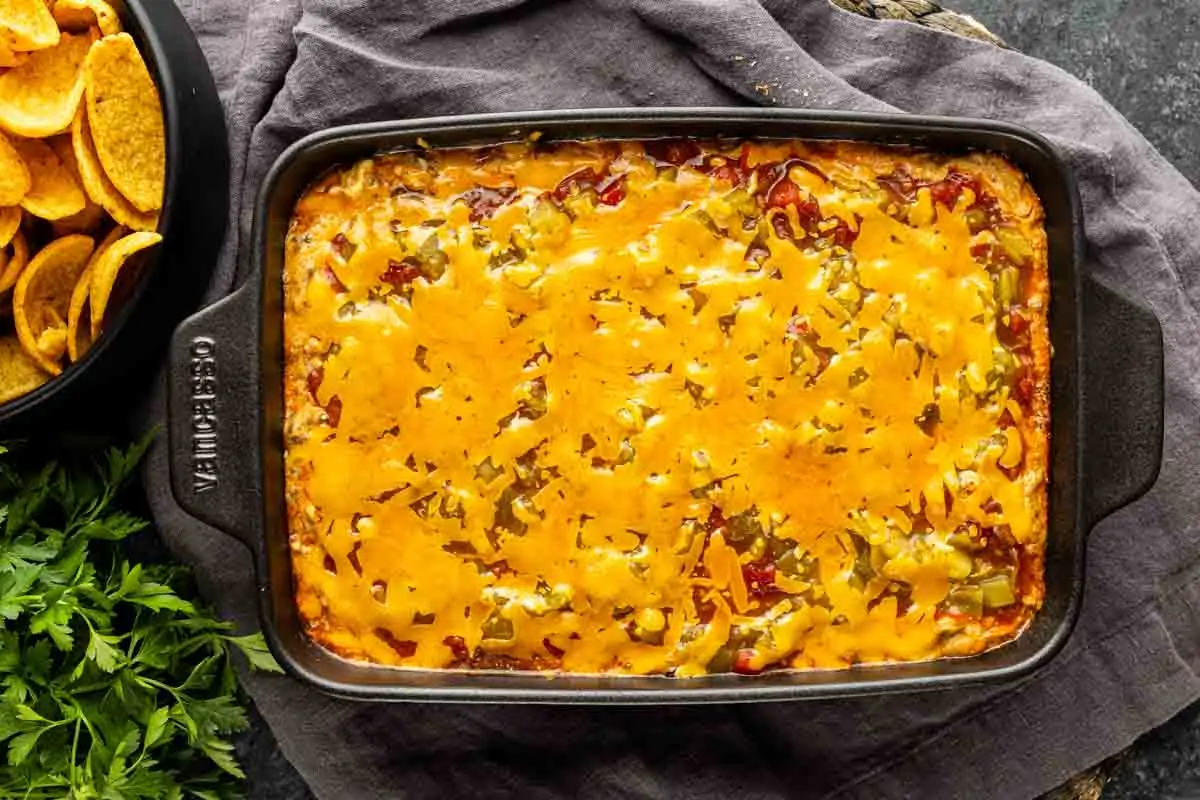 A casserole dish filled with baked cheesy taco dip with ground beef, topped with melted cheese and chopped peppers, sits next to a bowl of corn chips and fresh parsley.