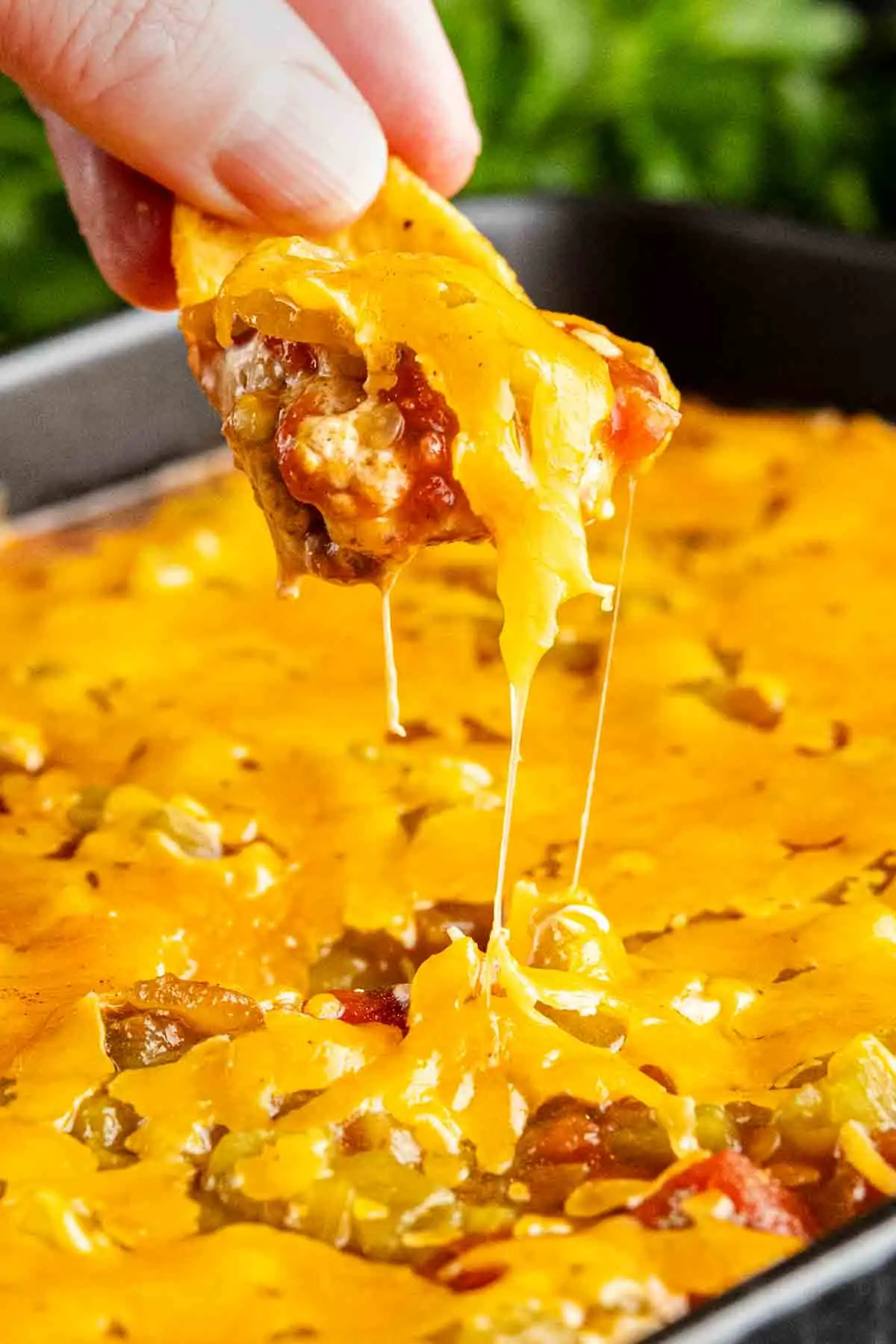 A hand dips a chip into a cheesy baked taco dip, lifting melted cheese and savory filling from the dish.