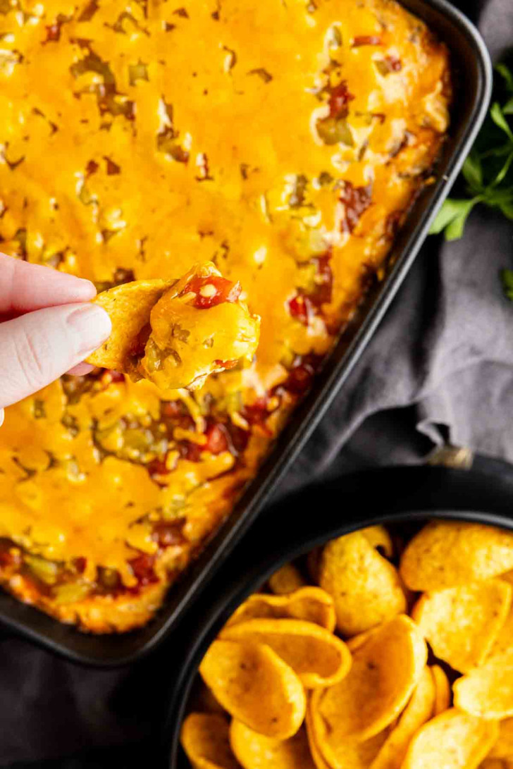 A hand dips a corn chip into a casserole dish filled with melted cheddar cheese dip—this easy taco dip with ground beef is served hot, with a bowl of crunchy corn chips nearby.