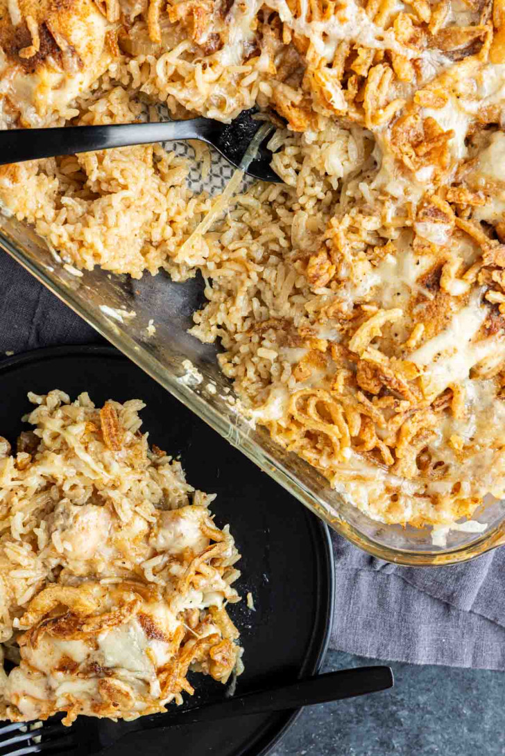 A casserole dish filled with a baked rice and chicken dish topped with crispy fried onions, with a serving on a black plate beside it.