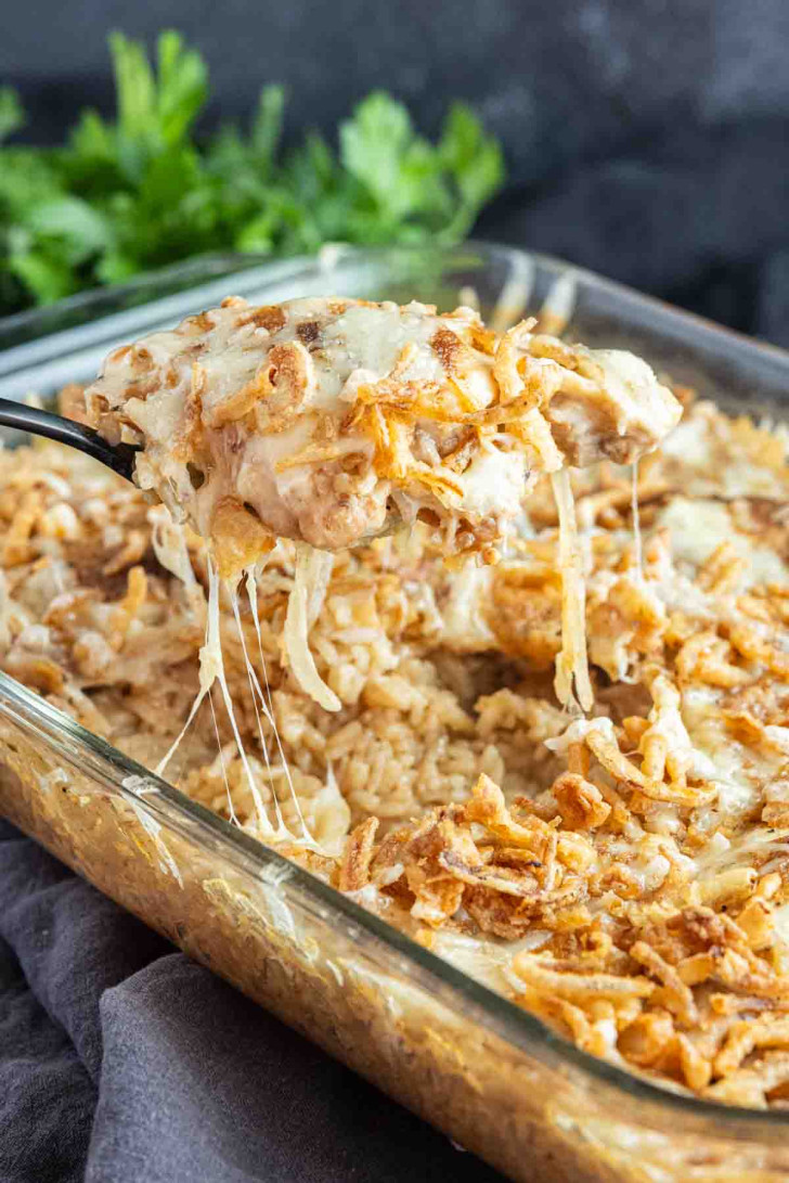 A casserole dish filled with a creamy, cheesy French Onion Chicken and Rice Bake topped with crispy fried onions; a serving is being lifted with a spoon.