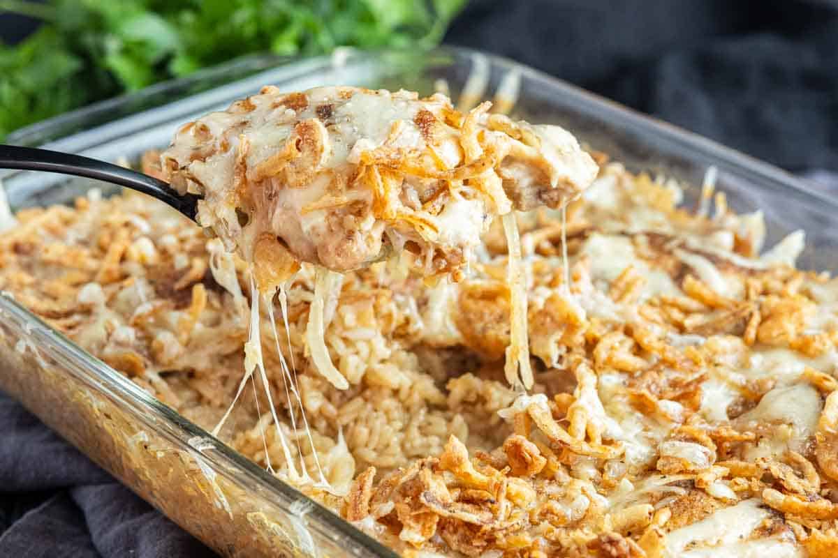 A serving spoon lifts a portion of cheesy, baked French Onion Chicken Bake with crispy fried onions from a glass dish.